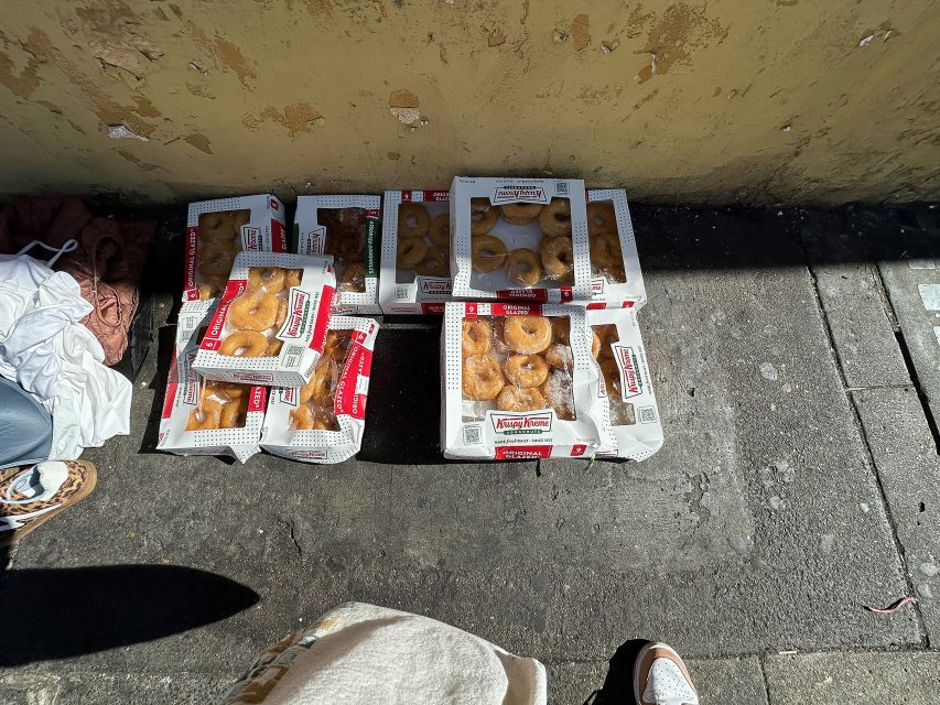 Several boxes of Krispy Kreme donuts are placed on a sunlit sidewalk next to a worn yellow wall, with part of a person’s foot and clothing visible at the edge.