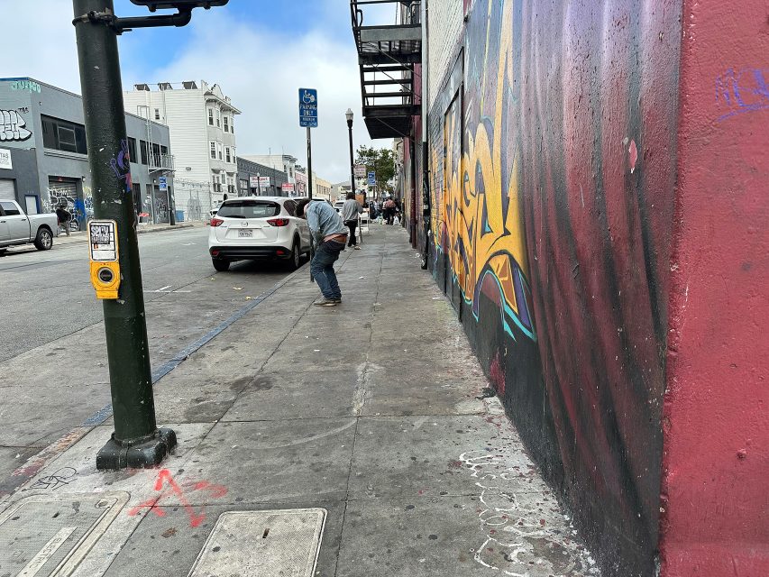 A man sweeps a gray city sidewalk next to a large mural, parked cars, and a traffic signal; buildings line the street under a partly cloudy sky.