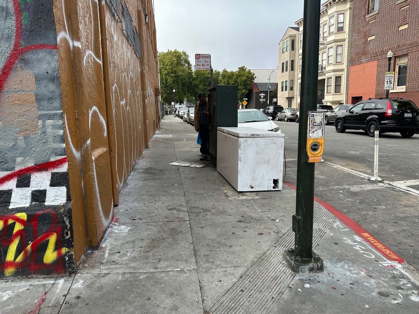 A city sidewalk with graffiti on the left wall, a large white appliance blocking part of the path, and buildings in the background.