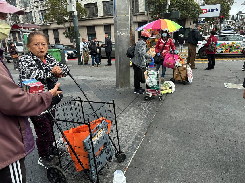 Several people with carts and bags gather on a city sidewalk near a street corner; one woman stands in the foreground, while others stand by an umbrella and bus stop.
