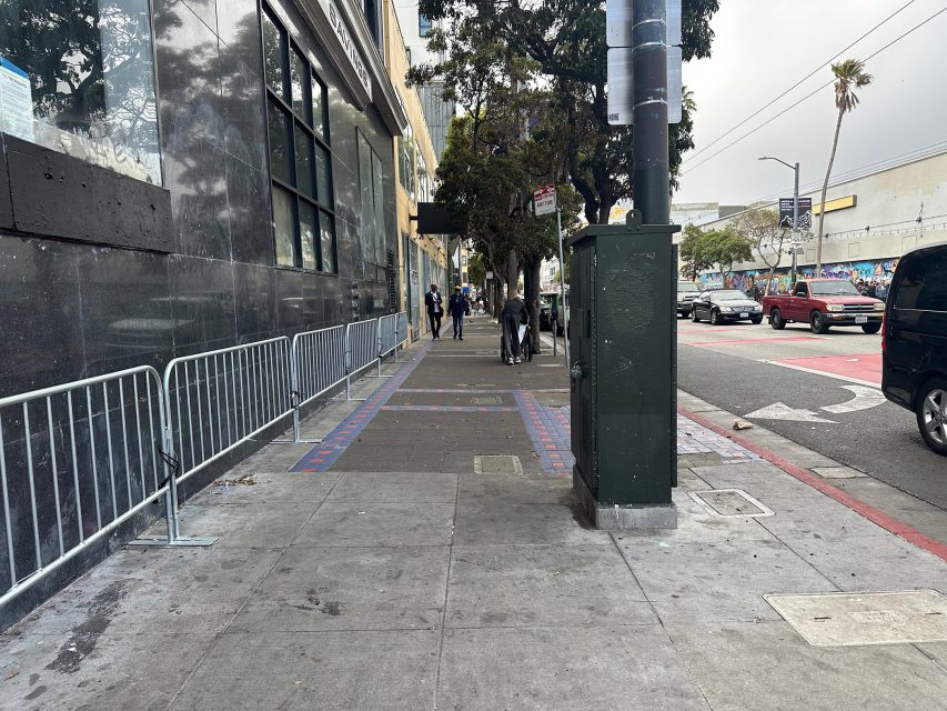 City sidewalk with metal barricades on the left, a utility box in the foreground, and people walking in the distance on an overcast day. Cars are parked and driving on the street.