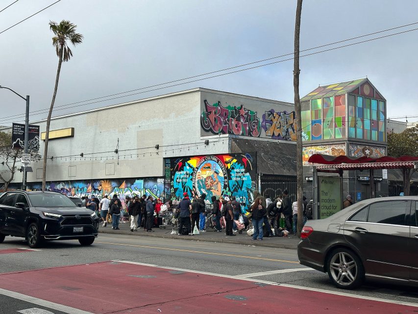 A long line of people waits outside a building with colorful murals on its walls, near a busy street with cars and a bus stop in the foreground.