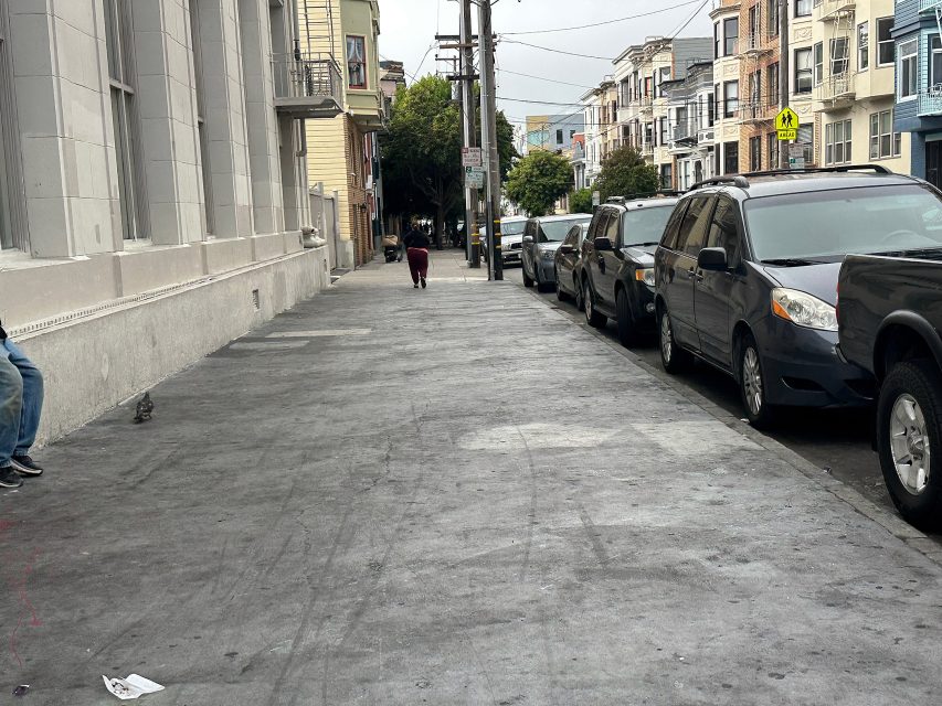 A mostly empty city sidewalk with parked cars on the right, a person walking away in the distance, and buildings lining both sides of the street.