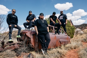 Five men dressed in black pose around and on top of a rusty, abandoned car in a dry, grassy outdoor setting with blue sky and scattered clouds.
