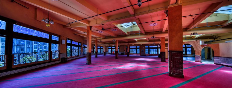 Spacious prayer hall with red carpet, large windows letting in natural light, and ornate architectural details on walls and ceiling.