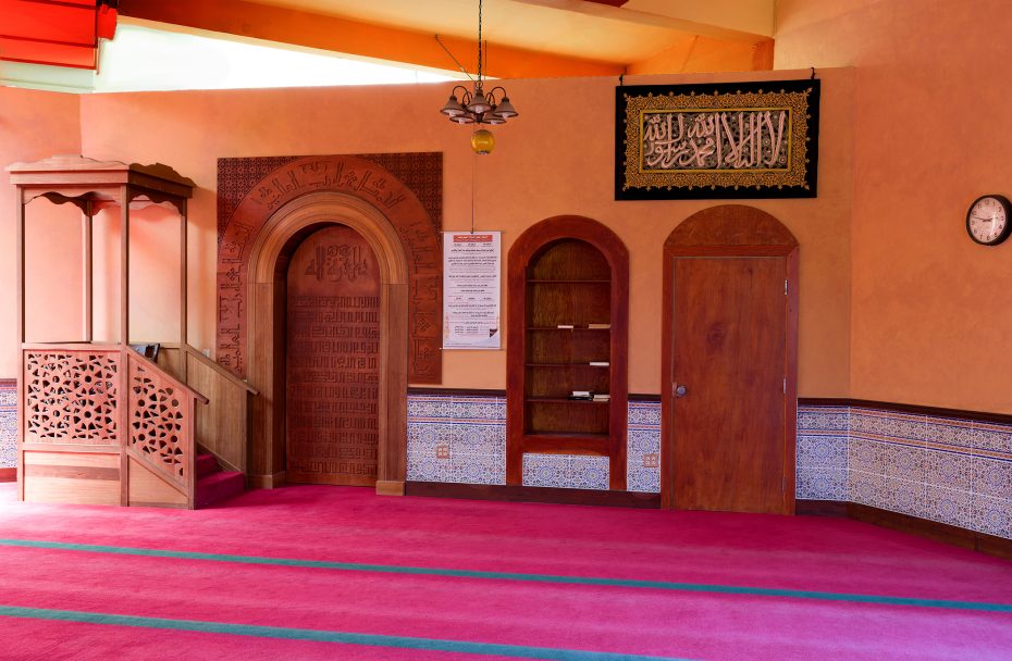 Interior of a mosque with red carpet, wooden mihrab and mimbar, decorative tiles, framed Arabic calligraphy, a wall clock, and a notice board.