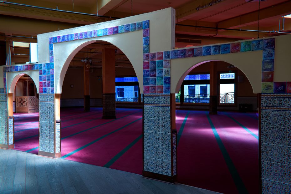 Three decorative arches with blue patterned tiles lead into a spacious prayer hall with red carpet and sunlight streaming through blue-tinted windows.