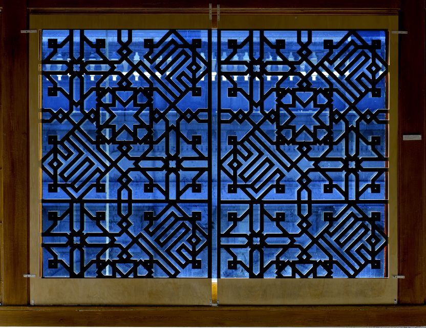 A wooden window screen features an intricate geometric lattice pattern, with blue light visible through the design.