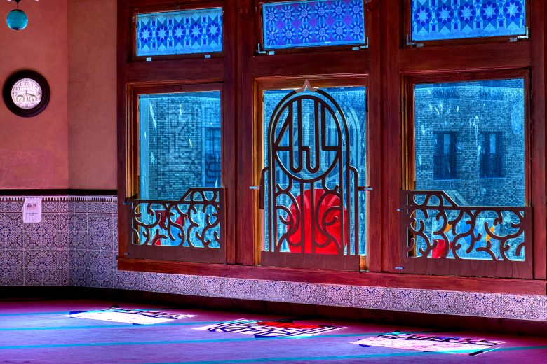 Interior of a mosque with patterned tiled walls, stained glass windows, and sunlight casting colorful patterns on the carpeted floor.