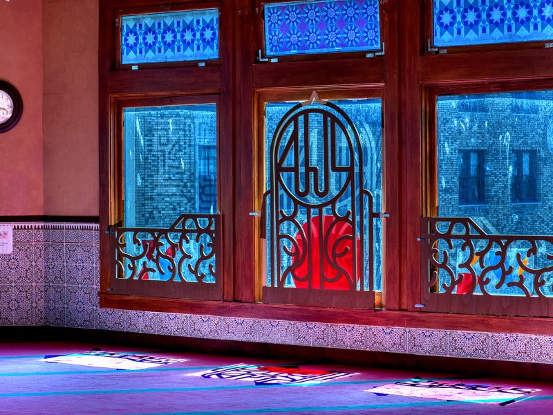 Interior of a mosque with patterned tiled walls, stained glass windows, and sunlight casting colorful patterns on the carpeted floor.