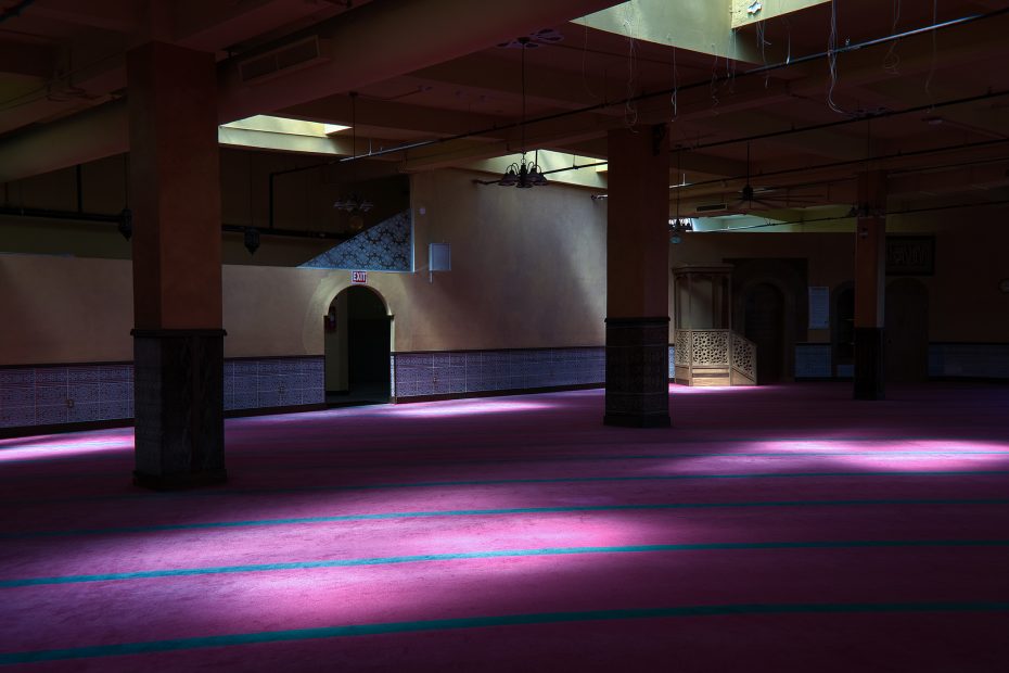 Sunlight streams through ceiling windows onto the pink carpet of a spacious, empty mosque interior, with columns, tiled walls, and an ornate mihrab visible in the background.
