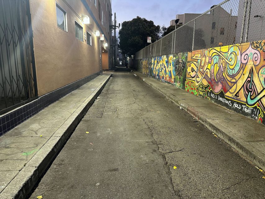 A narrow urban alley at dusk, bordered by a building on one side and a colorful graffiti-covered wall on the other, with a chain-link fence above the wall.