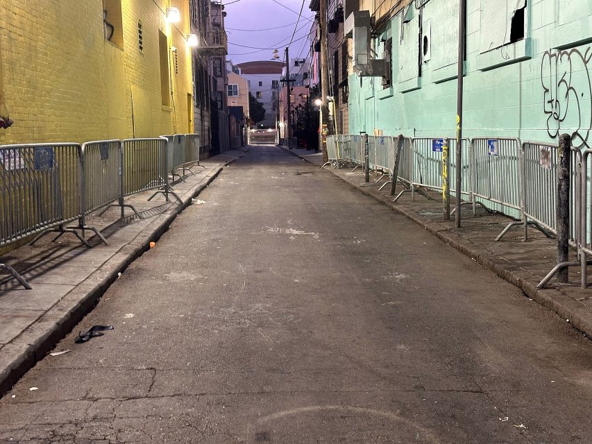 A deserted urban alleyway at dusk, lined with metal barricades and flanked by yellow and teal walls with graffiti and broken windows.