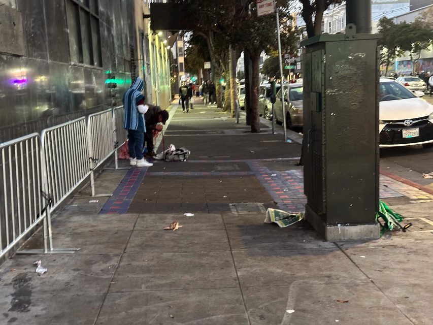 Two people stand on a city sidewalk near bags and belongings, with metal barricades and cars parked along the street. The scene appears cluttered and somewhat dirty.
