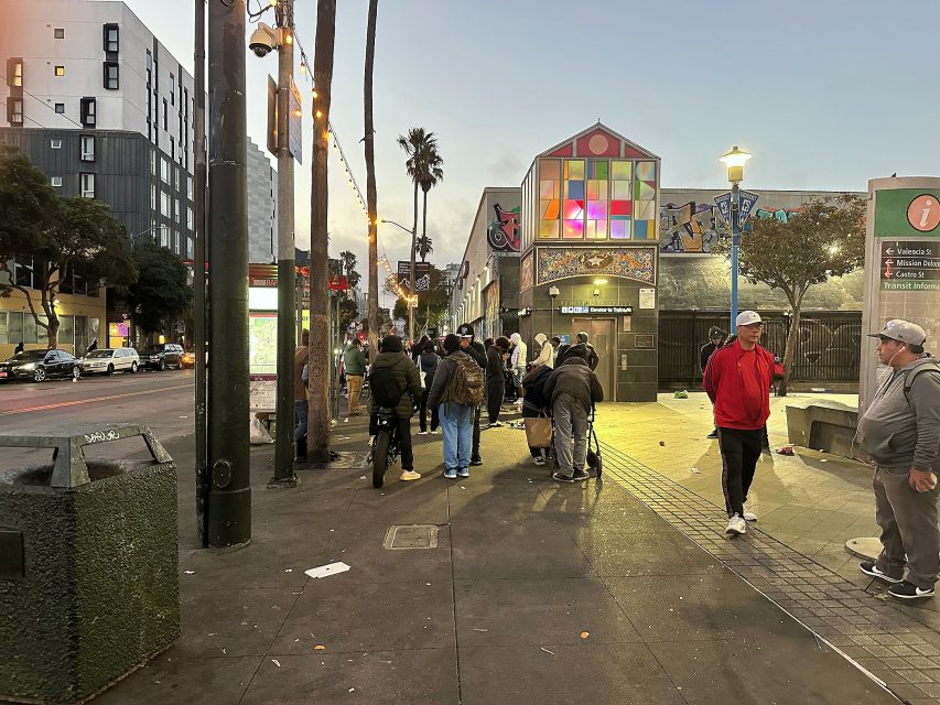 A group of people stand and gather on a city sidewalk near a colorful building at dusk, with trees, streetlights, and parked cars visible.