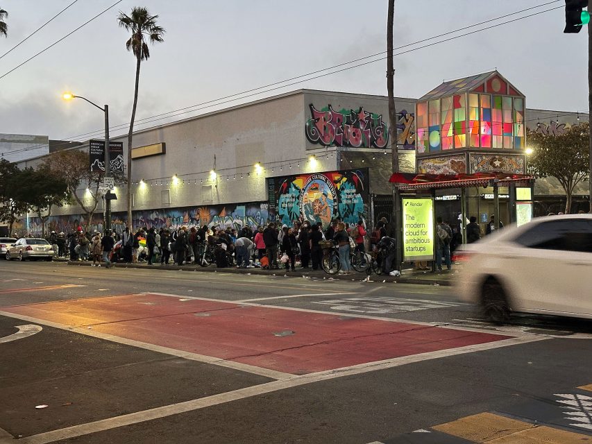 A long line of people stands outside a graffiti-covered building near a bus stop and a brightly lit glass structure, with cars passing by on the street.