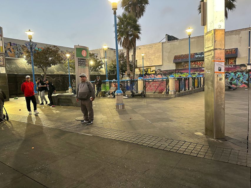 People standing and sitting in an urban plaza at dusk, surrounded by streetlights, palm trees, and walls covered in colorful graffiti.