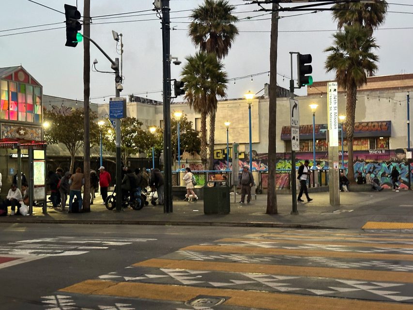Street scene at dusk with people waiting at a bus stop, palm trees, graffiti-covered walls, traffic lights, and crosswalk markings visible.