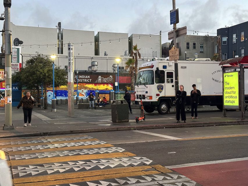 People stand at a street corner near a police vehicle and a yellow billboard reading "The modern cloud for ambitious startups" in an urban area at dusk.