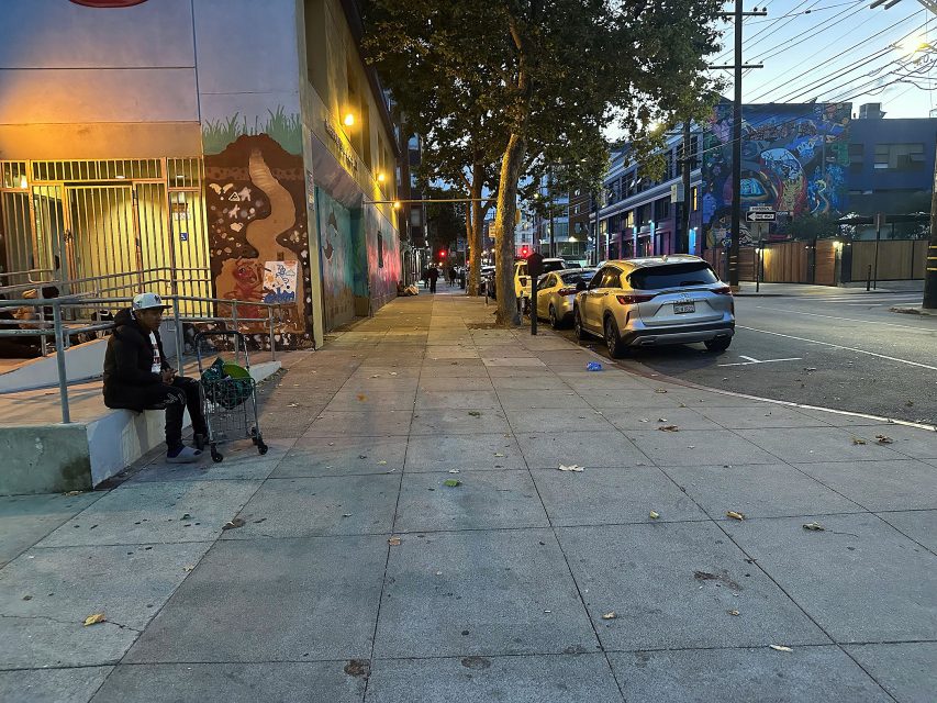 A person with a shopping cart sits on a concrete bench near a building with murals, as cars are parked along an empty city street at dusk.
