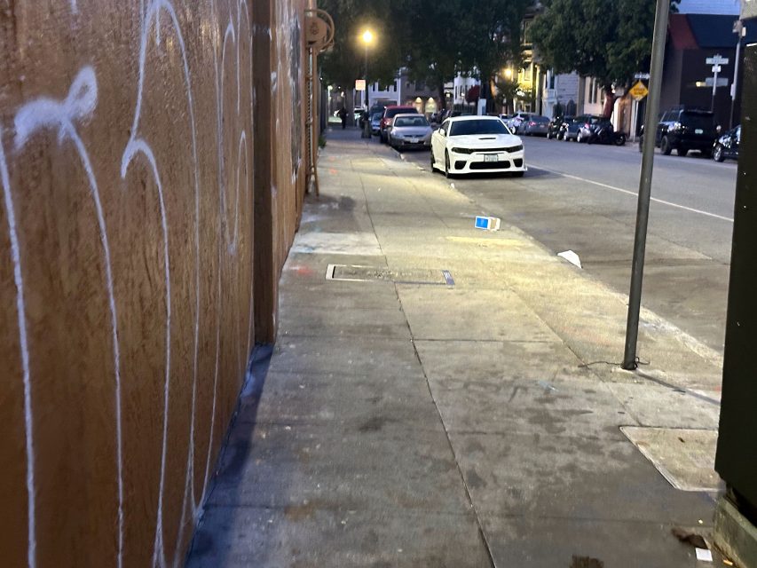 A city sidewalk at dusk with graffiti on the left wall, some litter on the ground, parked cars, and streetlights illuminating the scene.