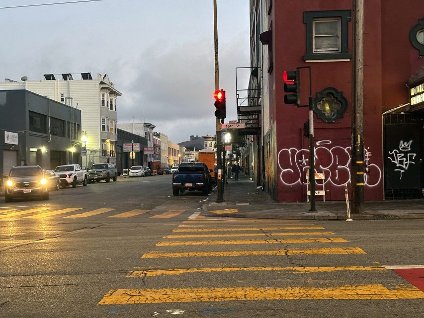 A city street intersection at dusk with a red traffic light, crosswalk lines, parked cars, and graffiti on a building wall.