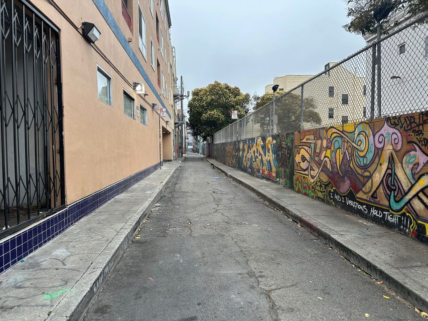 Narrow urban alleyway with graffiti on the right fence, a gated building on the left, and trees in the background under an overcast sky.