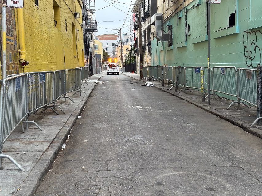 A narrow city alleyway with metal barricades on both sides, graffiti on walls, some litter on the ground, and an emergency vehicle with flashing lights in the distance.