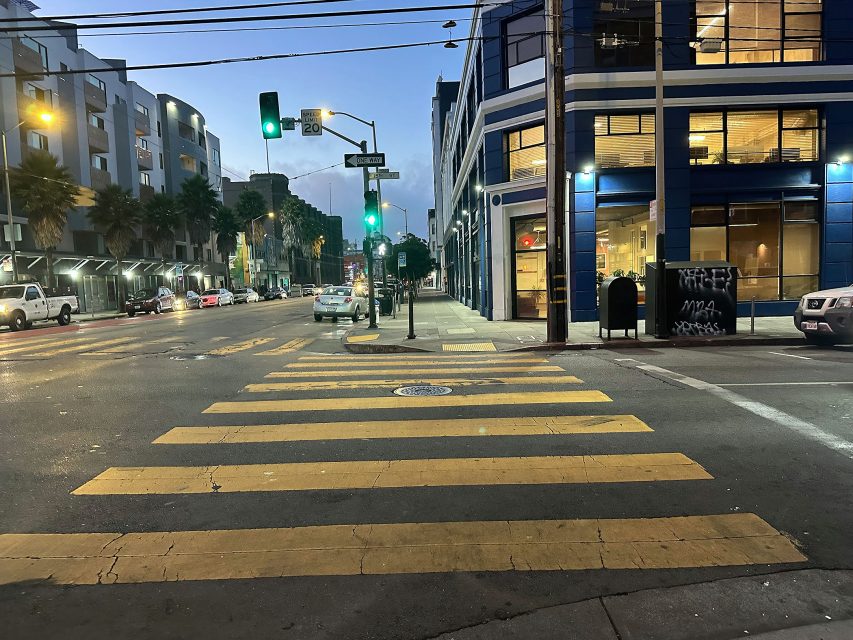 A city street intersection at dusk with a yellow crosswalk, traffic lights, and modern buildings on both sides.