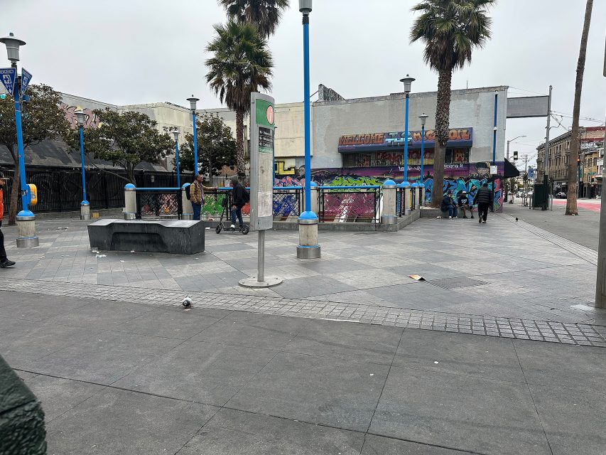 Urban plaza with blue lamp posts, palm trees, people walking, a black bench, and colorful graffiti on a building in the background. The sky is overcast.