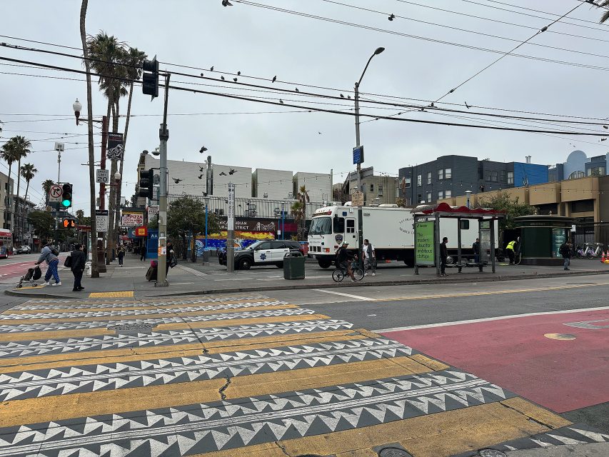 A white emergency response vehicle is parked near a bus stop on a busy city street with crosswalk markings and overhead power lines visible.