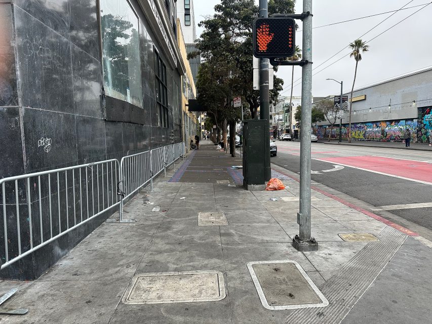 A city sidewalk with a pedestrian crossing signal showing a red hand, metal railings, scattered trash, and graffiti on a building across the street.