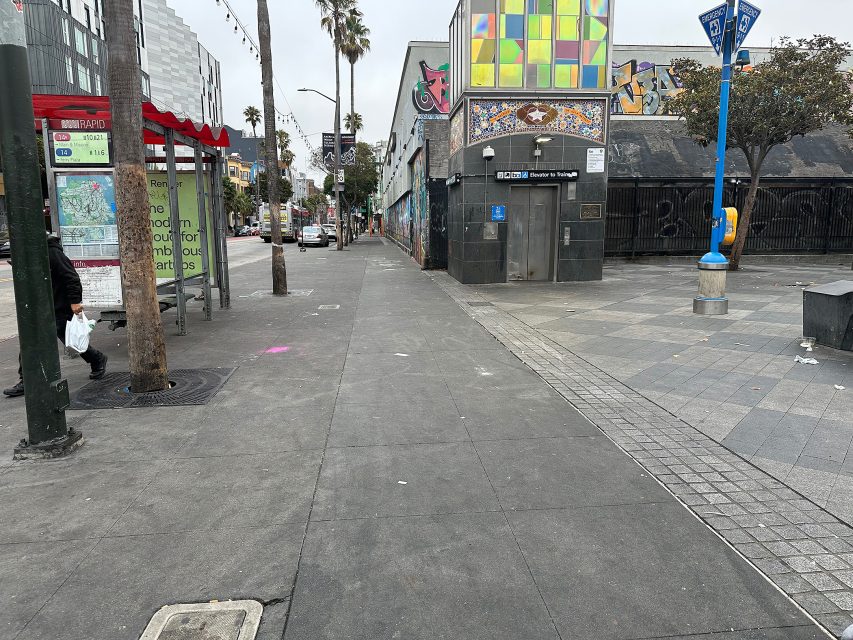 A mostly empty city sidewalk with a few trees, a red bus stop, some street art, and buildings on either side under an overcast sky.