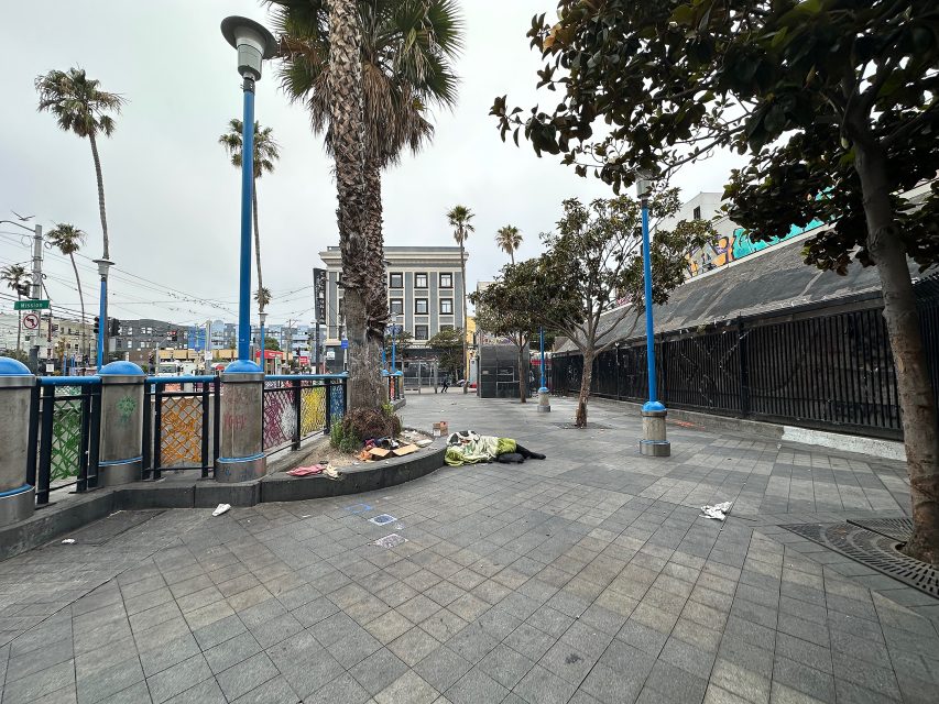 A mostly empty urban plaza with a few palm trees, scattered trash, and bedding on the ground beside a closed building on an overcast day.