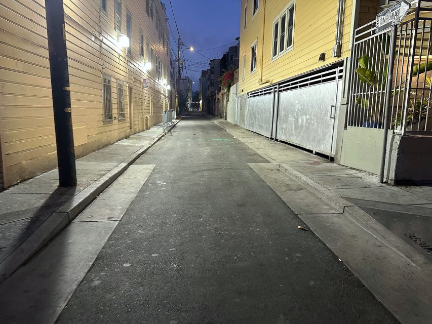 A narrow, empty alleyway between buildings at dusk, illuminated by several wall-mounted lights, with a gated area on the right side.