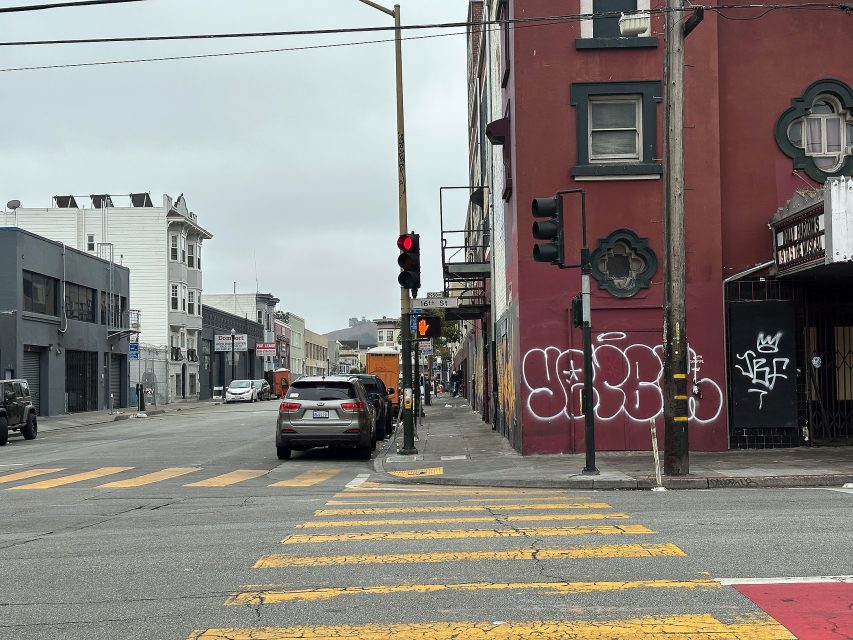 Urban street scene with cars parked along the curb, a red traffic light, crosswalk, and graffiti on a red building under an overcast sky.