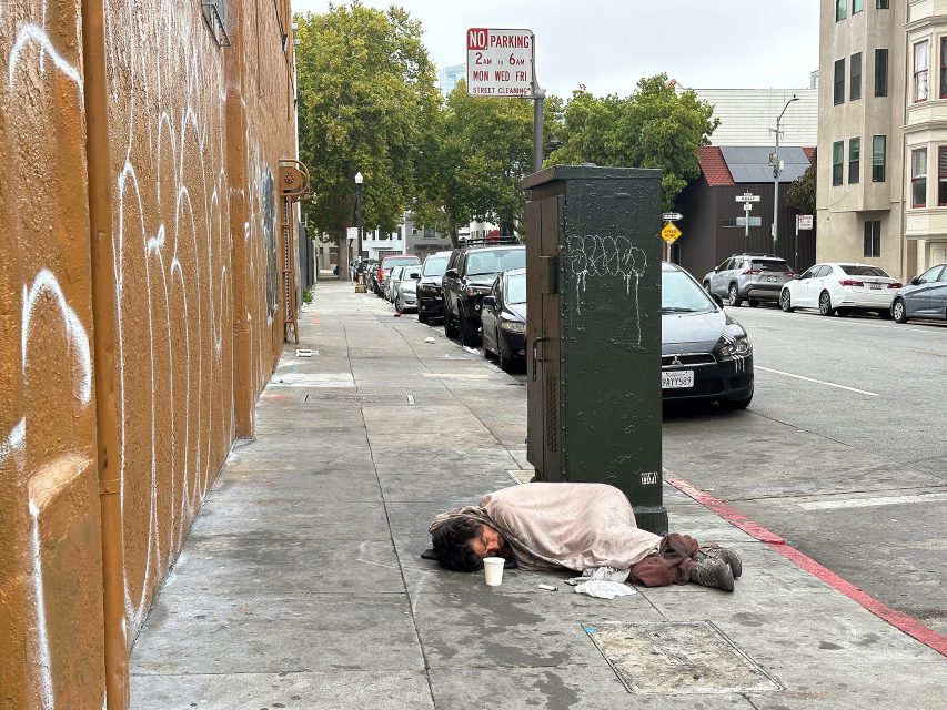 A person lies on a city sidewalk covered with a blanket next to a cup and trash, near parked cars and graffiti-covered walls.
