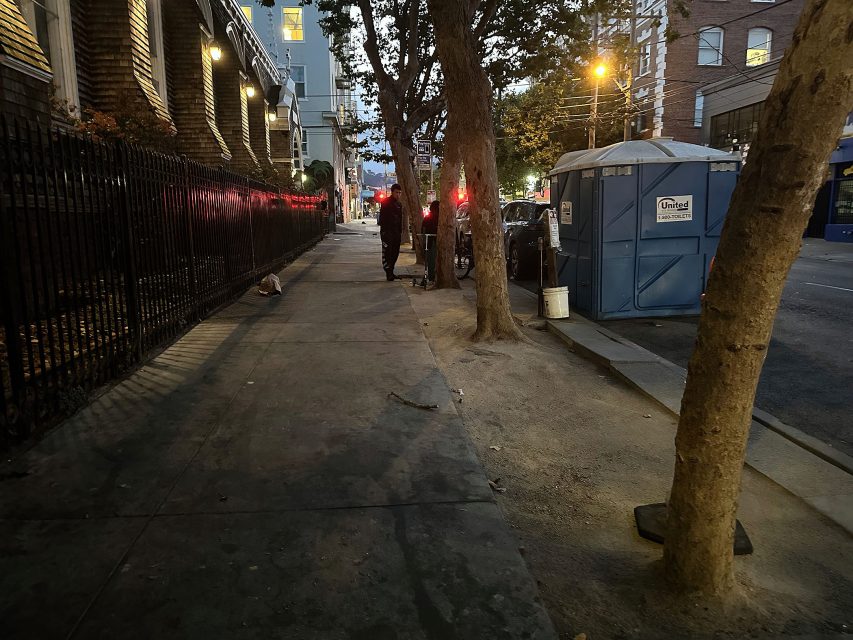 A person stands on a city sidewalk near a portable toilet and parked bicycles at dusk; trees and buildings line the street.