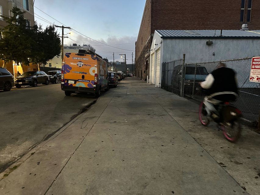 A person rides a bicycle on a city sidewalk at dusk; a parked food truck and cars line the street to the left.