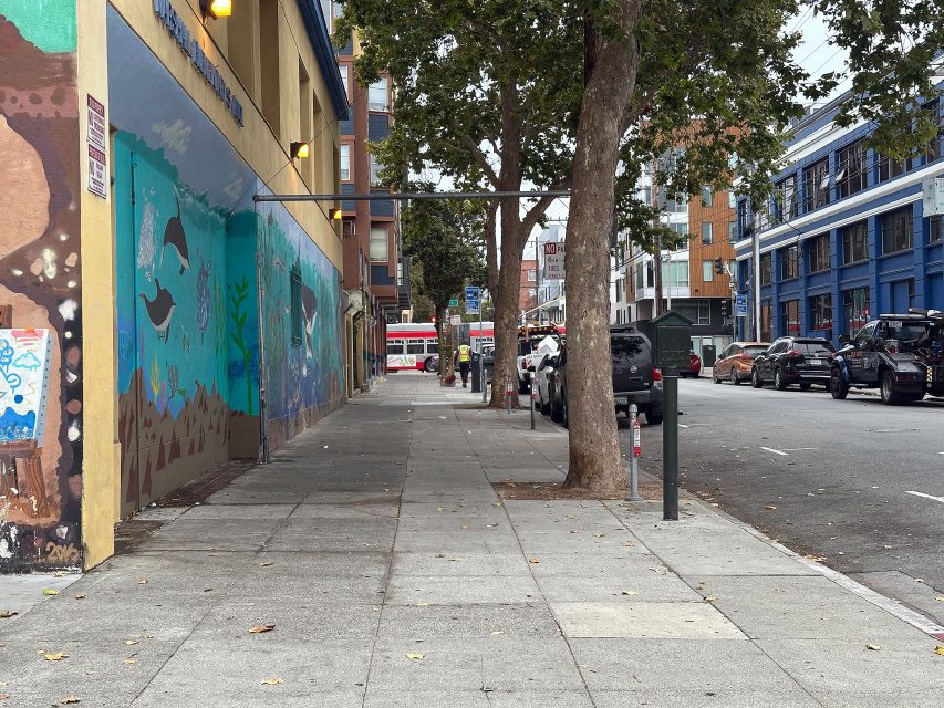 A city sidewalk lined with parked cars, trees, and a building featuring a colorful mural; a bus is visible in the distance on the street.