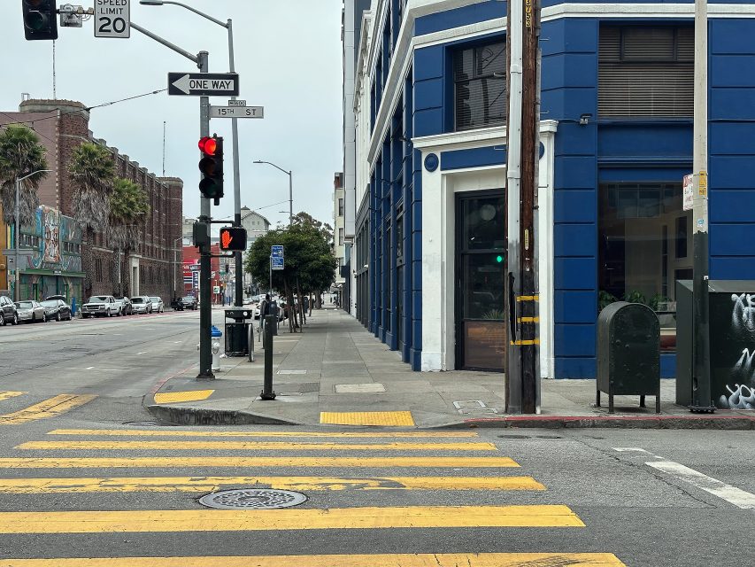 Empty city street with yellow crosswalk lines, traffic lights showing red and "Don't Walk" signal, and a blue building on the corner under an overcast sky.