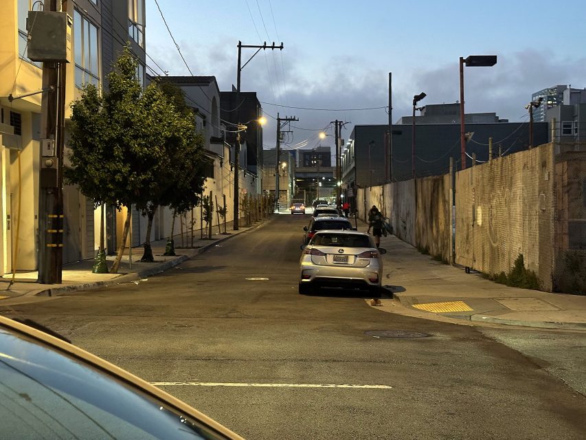 A quiet urban street at dusk with parked cars along both sides, a few people walking in the distance, and buildings lining the street.