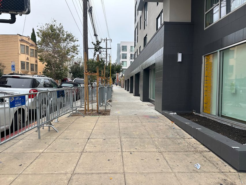 A city sidewalk next to a modern building, lined with parked cars, metal barricades, and power lines on a cloudy day.
