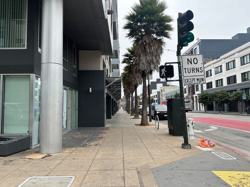 A city sidewalk with palm trees, modern buildings, a "No Turns Except Muni" sign, a traffic signal, and some trash bags near the curb.