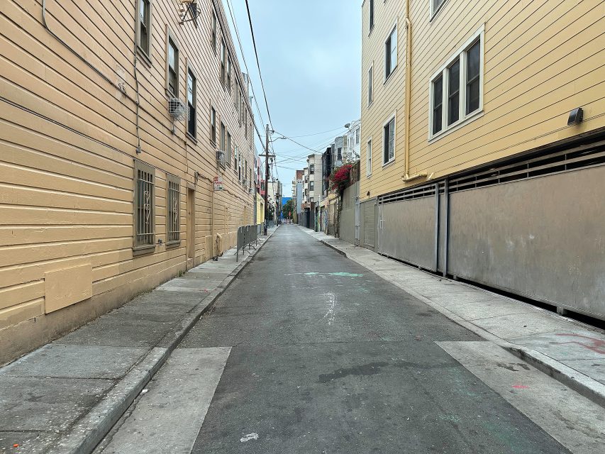 Narrow urban alleyway lined with beige and yellow buildings, overhead wires, and empty sidewalks on a cloudy day.