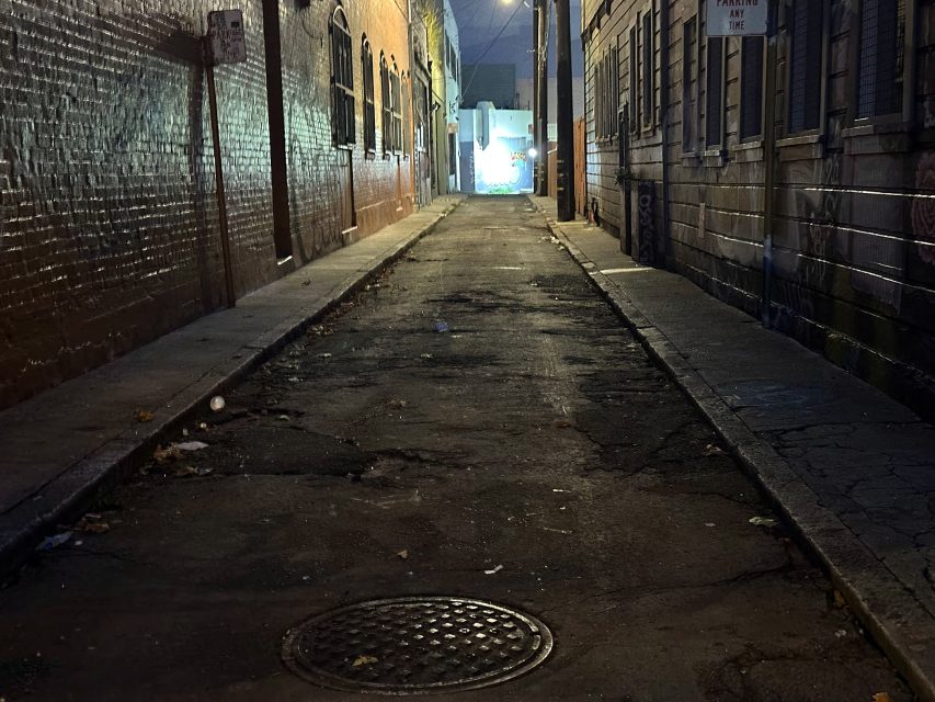 A narrow, dimly lit alleyway with cracked pavement, bordered by brick and wood buildings, leading to a brightly lit area in the distance.