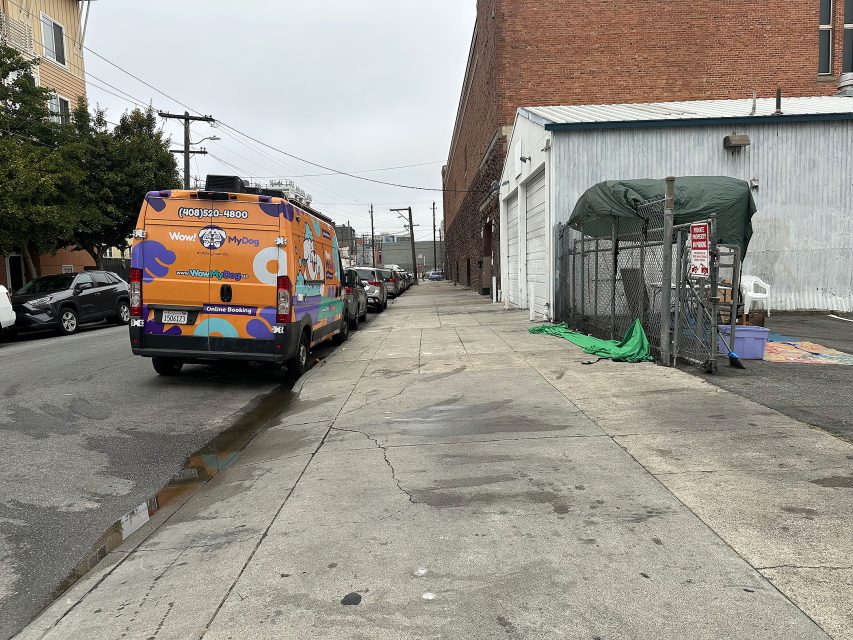 A colorful van is parked on a city street beside a sidewalk, near a fenced area with a tarp and a metal building; overcast sky overhead.