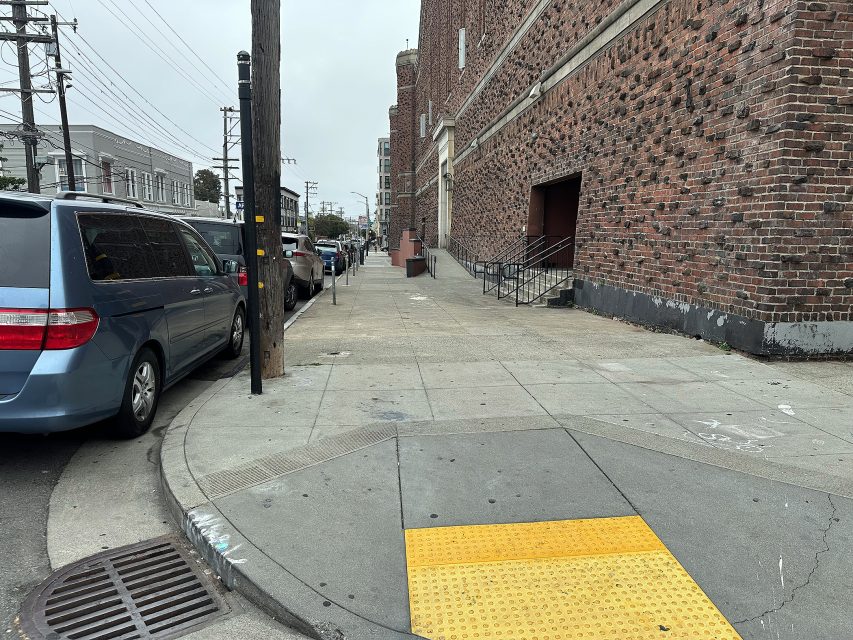 Sidewalk with curb ramp and yellow tactile paving beside a brick building; parked cars line the street under overcast skies.