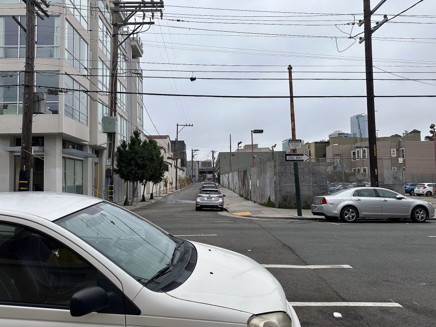 A city street intersection with parked cars, buildings on the left, chain-link fences on the right, and utility poles with overhead wires on a cloudy day.