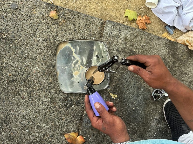 Two hands hold tools to cut a lock submerged in a bowl of murky water on a concrete surface with scattered debris and leaves.